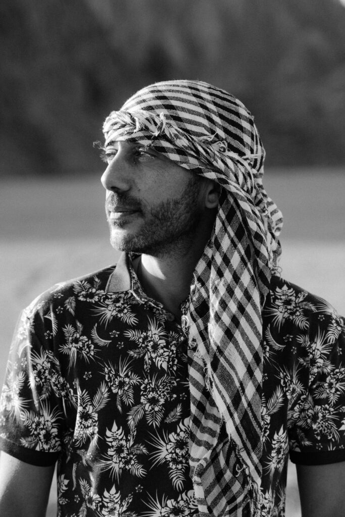 Black and white portrait of a bearded man wearing a traditional headscarf outdoors.