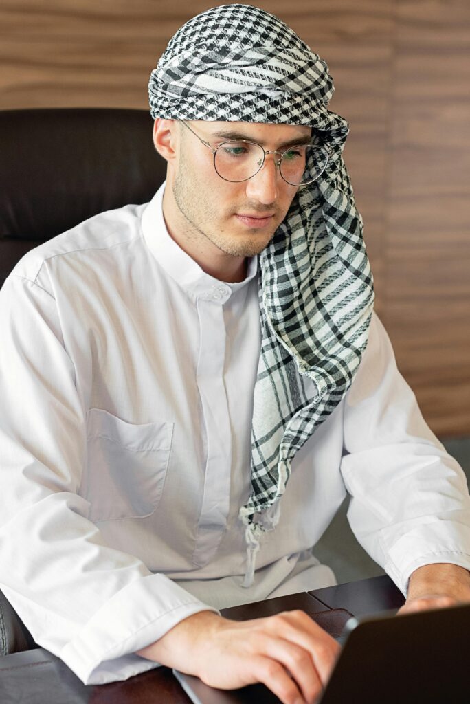 A focused Middle Eastern man with traditional attire and glasses working on a laptop indoors.